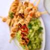 A close up of a grilled shrimp skewer seasoned with Jr Lemon Pepper Seas X6 and garnished with a red chili slice held over a plate of fresh cucumber salad.