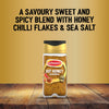 A bottle of Schwartz Jr Hot Honey Seas X6 stands on a rustic wooden surface against a golden background featuring text describing the savoury sweet and spicy blend with honey chilli flakes and sea salt.