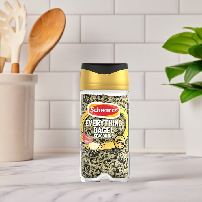 A bottle of Schwartz Jr Every Bagel Seas X6 stands on a marble countertop in a bright kitchen setting with cooking utensils and a plant in the background.