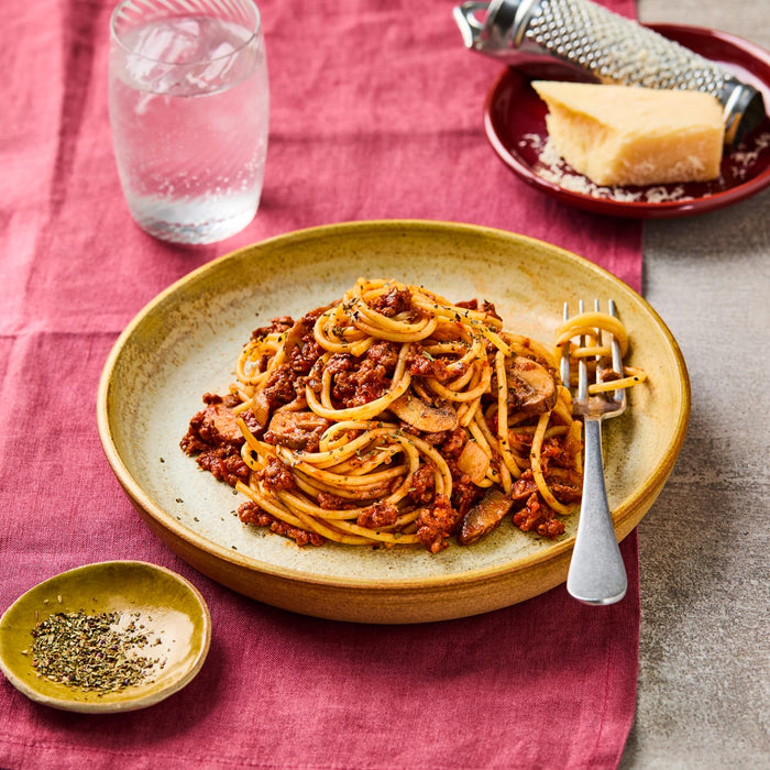 A plate of spaghetti bolognese is garnished with a sprinkle of Schwartz Italian Herb Seasoning 11G alongside a wedge of parmesan cheese and a glass of water.