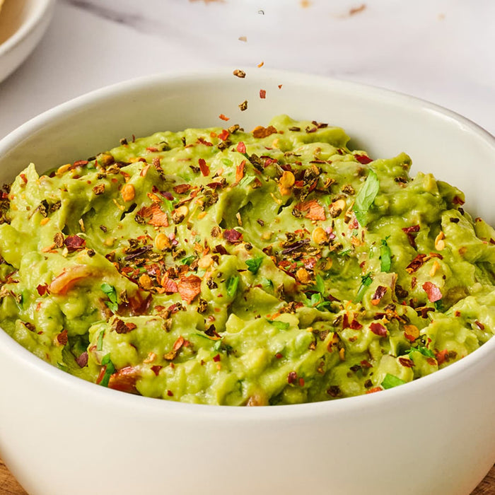 A close-up shot shows a white bowl of chunky guacamole being topped with a sprinkle of Schwartz Crushed Chillies 29G for a spicy kick.