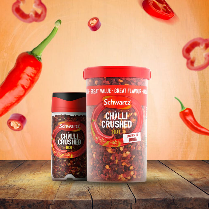 A small jar of Schwartz Crushed Chillies 29G and a larger value tub are displayed on a rustic wooden table, with fresh and sliced red chillies in the background to showcase the main ingredient.