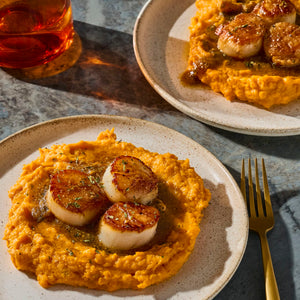 Two plates of seared scallops served on a bed of mashed sweet potatoes or squash, garnished with herbs and a sauce, a meal that could incorporate the rich flavor of Mccormick Gourmet Nutmeg Whole.