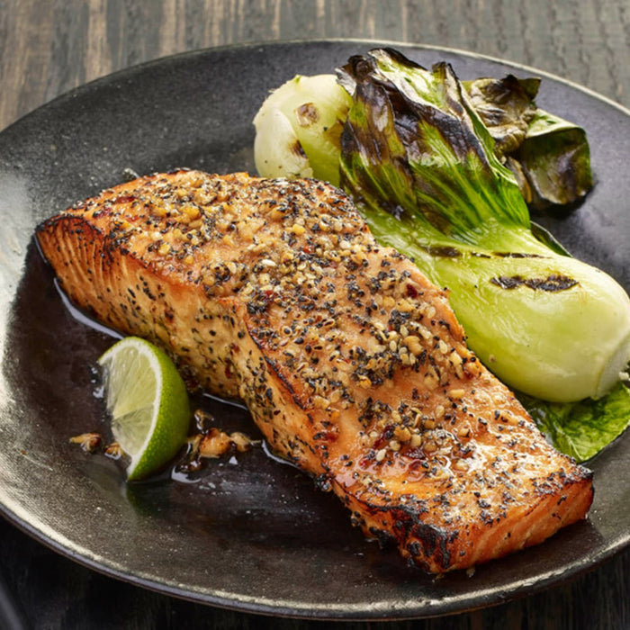 A delicious-looking salmon fillet, seasoned with Schwartz Ground Black Pepper, 33G, is served on a dark plate with grilled bok choy and a lime wedge.