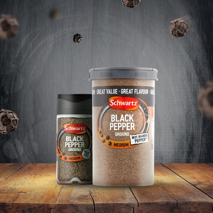 A smaller shaker and a larger tub of Schwartz Ground Black Pepper, 33G, are displayed on a rustic wooden table with whole peppercorns floating in the background.