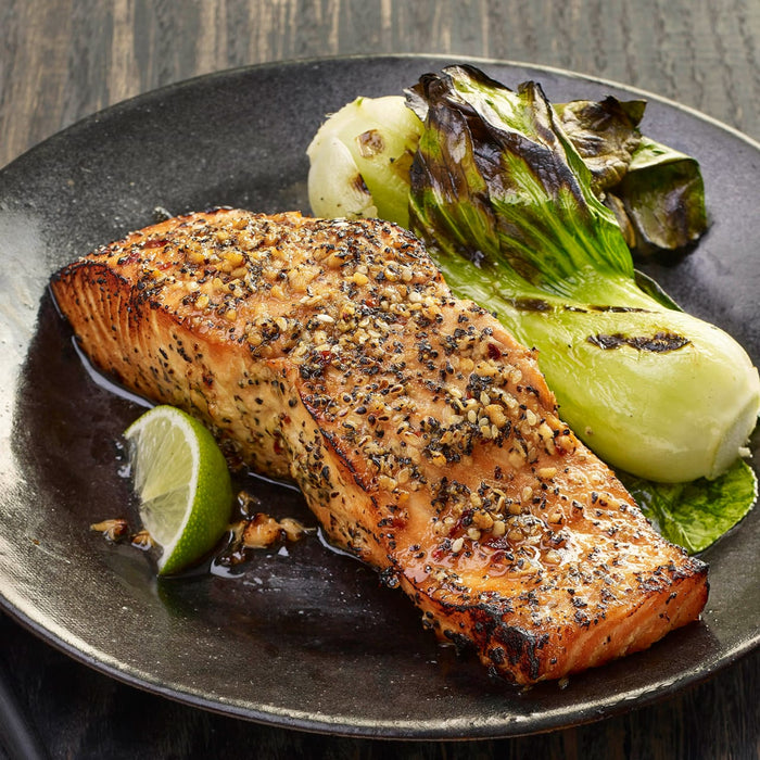 A perfectly seared salmon fillet seasoned with a savory spice blend is served on a dark plate alongside grilled bok choy and a fresh lime wedge.