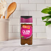 A bottle of Schwartz Cajun Seasoning 44G stands on a marble countertop in a bright kitchen setting with wooden utensils and a green plant in the background.