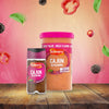 A small jar and a large catering tub of Schwartz Cajun Seasoning 44G are displayed on a wooden table with floating ingredients like peppers and leaves against a pink background.