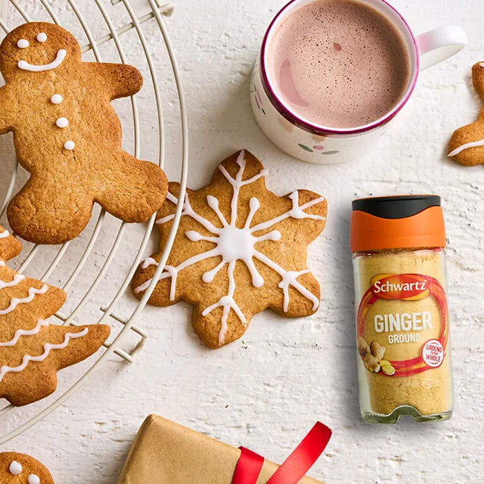 A festive holiday scene featuring decorated gingerbread cookies and a mug of hot cocoa alongside a Schwartz Ground Ginger 26G Jar used for baking.