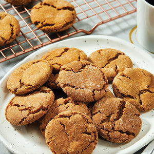 A plate of warm ginger snap cookies made with Schwartz Ground Ginger 26G Jar sits next to a cooling rack and a cup of coffee.