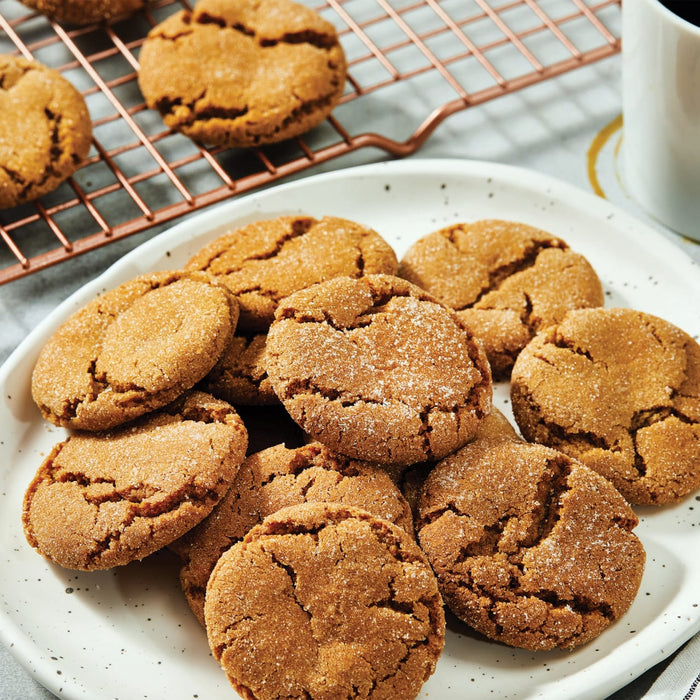A plate of warm ginger snap cookies made with Schwartz Ground Ginger 26G Jar sits next to a cooling rack and a cup of coffee.