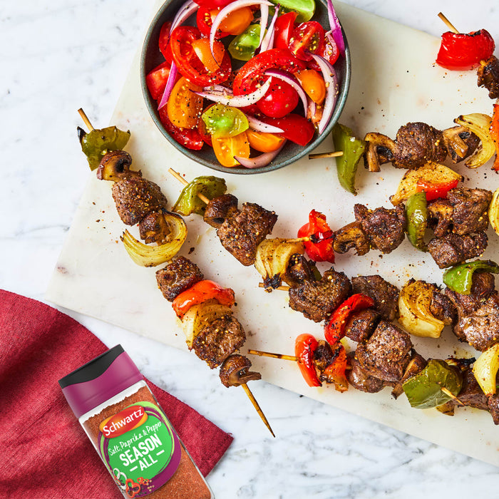 A bottle of Schwartz Salt, Paprika & Pepper Season All Seasoning 70G sits next to a platter of grilled beef and vegetable skewers served with a fresh tomato and onion salad.