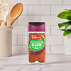 A bottle of Schwartz Salt, Paprika & Pepper Season All Seasoning 70G stands on a marble countertop in a bright kitchen setting with cooking utensils and a green plant in the background.