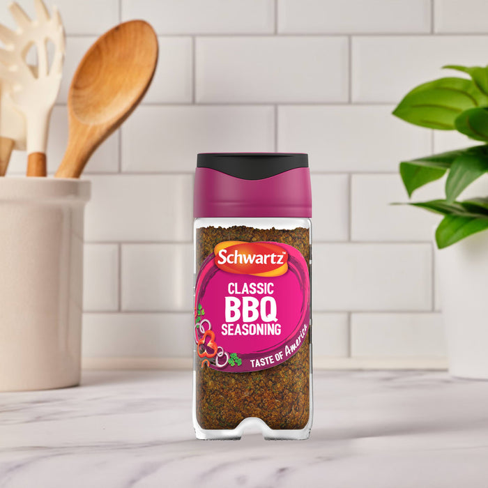 A bottle of Schwartz Classic Bbq Seasoning 44G stands on a marble countertop in a bright kitchen setting with wooden utensils and a green plant in the background.
