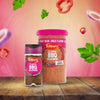 A small jar and a large catering tub of Schwartz Classic Bbq Seasoning 44G are displayed on a wooden surface against a pink background with floating ingredients like red peppers and onions.