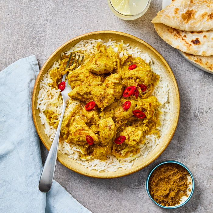 A delicious-looking bowl of yellow chicken curry is served over a bed of white rice, garnished with red chili slices, and accompanied by flatbread and a small bowl of curry powder.