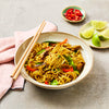 A rustic bowl filled with stir-fried noodles, chicken, and vegetables, served with chopsticks and a side of fresh lime and chili.