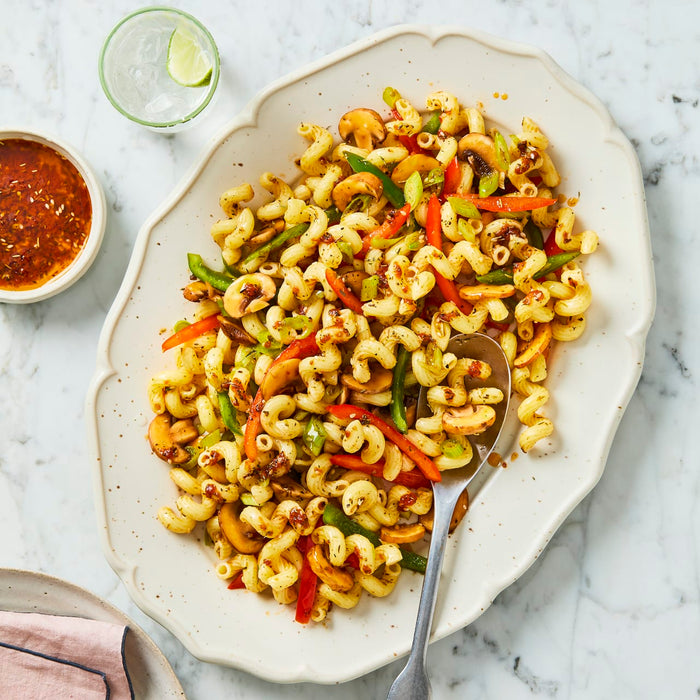 A large, decorative white platter of spiral pasta salad with mushrooms and peppers is shown on a marble countertop, with a side of dressing and a glass of water.