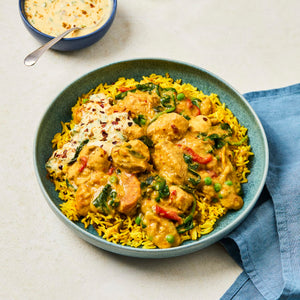 A delicious-looking chicken curry with vegetables is served over a bed of yellow rice in a blue bowl, with a side of creamy sauce.