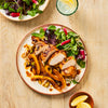 An overhead view of a plated meal featuring sliced, glazed chicken breast, cooked peppers, and a side salad with radishes and watercress.