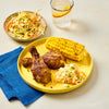 A yellow plate holds two pieces of seasoned chicken, corn on the cob, and a side of coleslaw, with a separate bowl of coleslaw and a drink in the background.