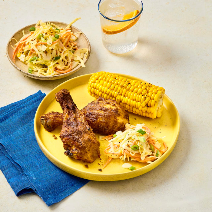 A yellow plate holds two pieces of seasoned chicken, corn on the cob, and a side of coleslaw, with a separate bowl of coleslaw and a drink in the background.