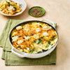 A freshly baked pasta casserole with spinach and cheese sits in a white dish, with a serving on a plate and a small dish of coarsely ground black pepper nearby.