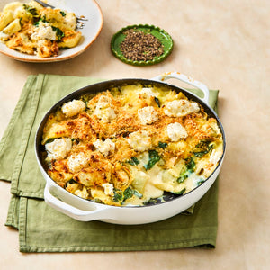 A freshly baked pasta casserole with spinach and cheese sits in a white dish, with a serving on a plate and a small dish of coarsely ground black pepper nearby.