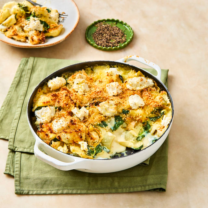 A freshly baked pasta casserole with spinach and cheese sits in a white dish, with a serving on a plate and a small dish of coarsely ground black pepper nearby.
