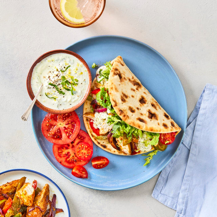A freshly prepared chicken gyro is presented on a blue plate with a side of tzatziki sauce, sliced tomatoes, and a glass of sparkling water with lemon.