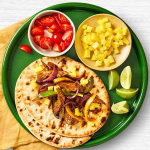 A vibrant green plate holds two flatbreads topped with seasoned meat, onions, and peppers, with side bowls of diced pineapple and tomatoes, and fresh lime wedges.