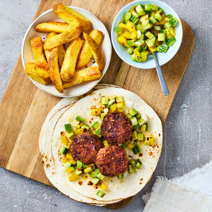 A top-down view of a meal on a wooden board, featuring patties on tortillas with a fresh salsa, a side of seasoned potato wedges, and a bowl of cucumber and mango salsa.