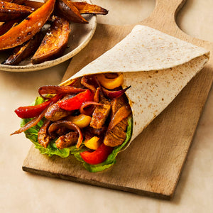 A freshly made fajita wrap filled with seasoned meat, peppers, and onions sits on a wooden board next to a bowl of sweet potato wedges.