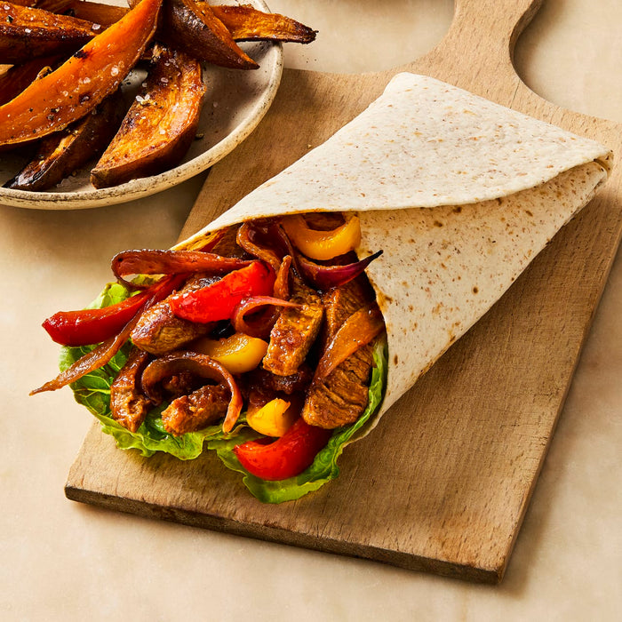 A freshly made fajita wrap filled with seasoned meat, peppers, and onions sits on a wooden board next to a bowl of sweet potato wedges.