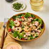 A rustic brown bowl is filled with a shrimp and rice salad, drizzled with a creamy pink dressing and garnished with fresh herbs.