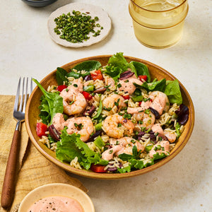 A rustic brown bowl is filled with a shrimp and rice salad, drizzled with a creamy pink dressing and garnished with fresh herbs.