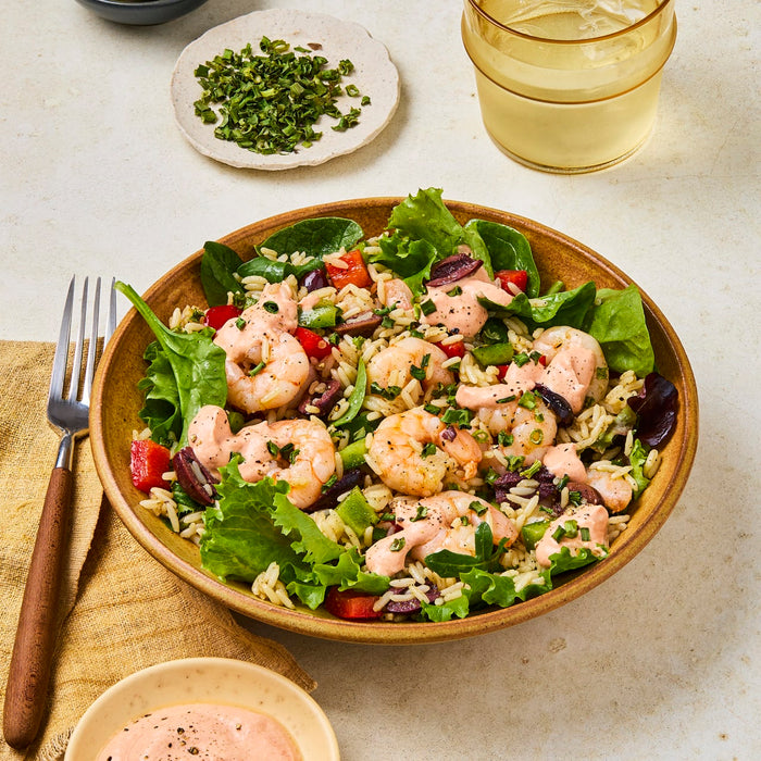 A rustic brown bowl is filled with a shrimp and rice salad, drizzled with a creamy pink dressing and garnished with fresh herbs.