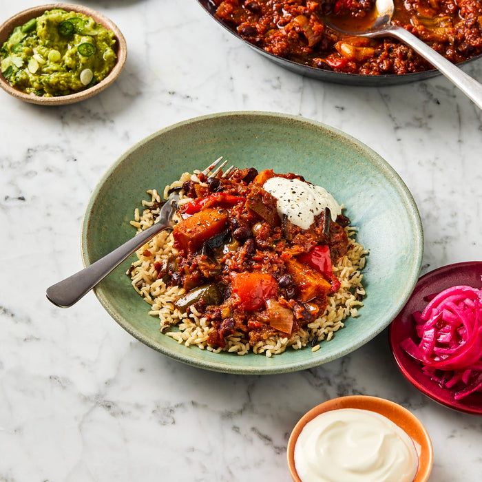 A bowl of vegetarian chili is served over brown rice with a dollop of sour cream, alongside small bowls of guacamole and pickled onions.