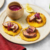 Three small appetizers of seared duck breast and a rich berry sauce are served on golden corn cakes, garnished with lemon zest on a rustic plate.