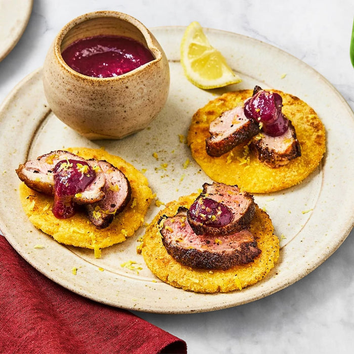 Three small appetizers of seared duck breast and a rich berry sauce are served on golden corn cakes, garnished with lemon zest on a rustic plate.