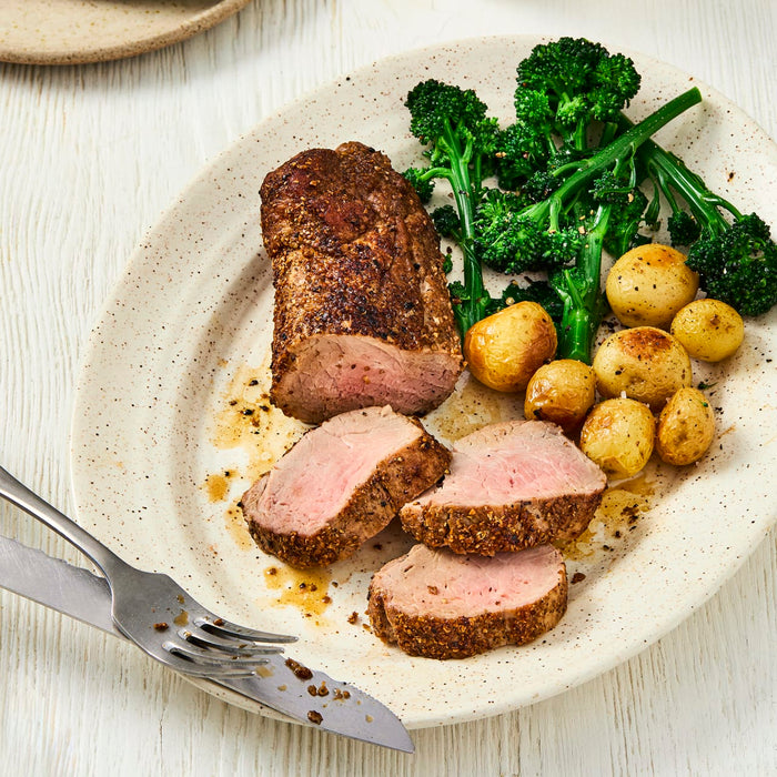 A sliced, spice-crusted pork tenderloin is plated with roasted new potatoes and broccolini, with a fork and knife resting on the side.