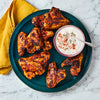 A dark green plate holds several grilled chicken wings next to a small bowl of creamy blue cheese dipping sauce, all set on a white marble countertop.