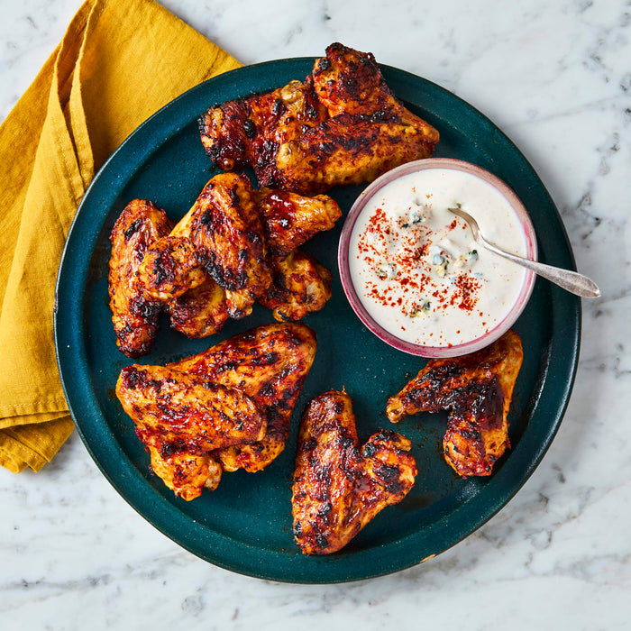A dark green plate holds several grilled chicken wings next to a small bowl of creamy blue cheese dipping sauce, all set on a white marble countertop.