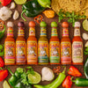 A variety of Cholula hot sauce bottles are arranged with peppers, limes, garlic, and cilantro.