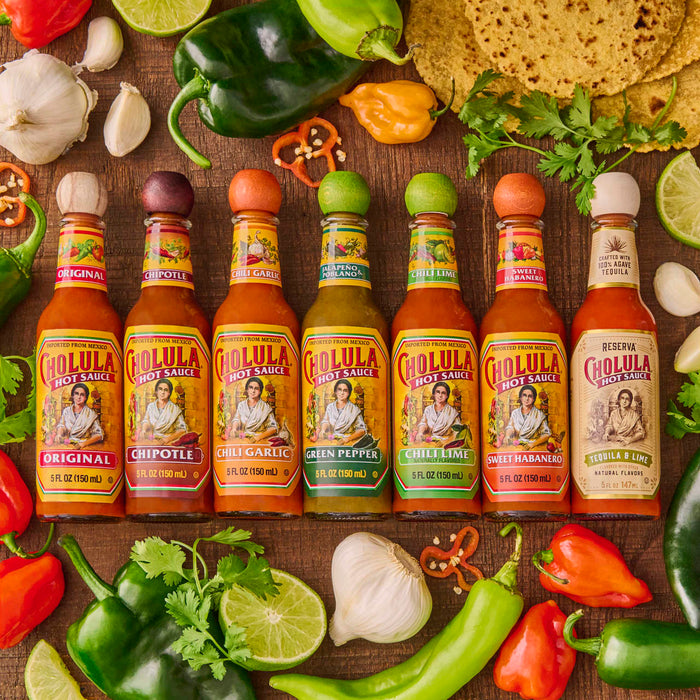 A variety of Cholula hot sauce bottles are arranged with peppers, limes, garlic, and cilantro.