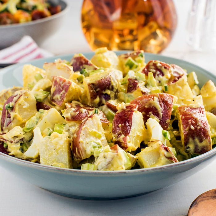 A light blue bowl is filled with a creamy red potato salad, with a large glass Barrel drink dispenser and another salad in the background.