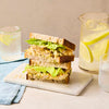 A delicious-looking coronation chicken salad sandwich is cut in half and stacked on a marble board, with a pitcher of lemon water in the background.