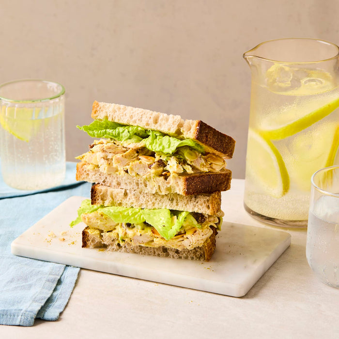 A delicious-looking coronation chicken salad sandwich is cut in half and stacked on a marble board, with a pitcher of lemon water in the background.