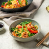A rustic Barrel bowl is filled with a vibrant shrimp and noodle stir-fry, with a wok of the same dish visible in the background.