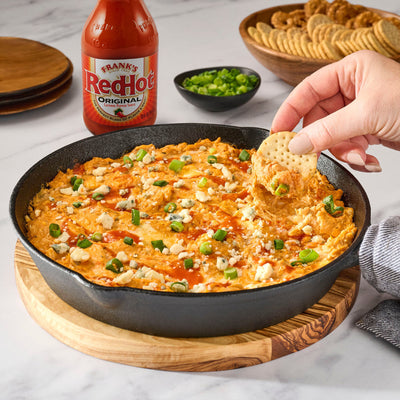 Hot sauce bottle, bowl of green onions, and hand dipping a cracker into buffalo chicken dip.